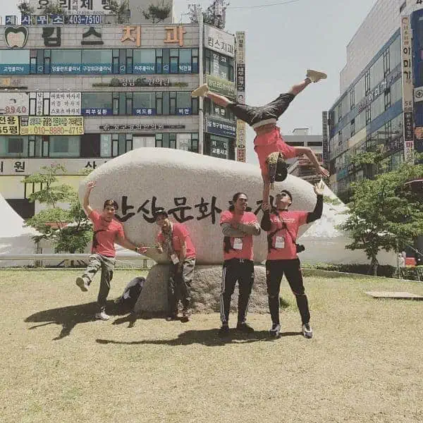 Phare Circus performers outdoors in Korea, one-handed partner handstand. Others waiving