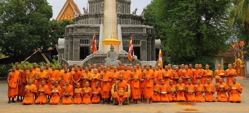 Group of monks in orange robes holding candles, seated and standing in front of a pagoda