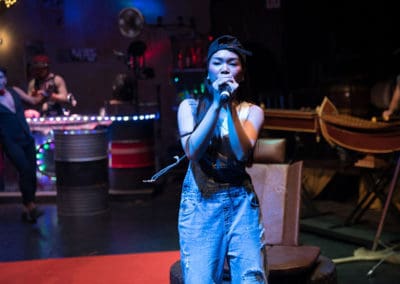 Phare Circus performance "Khmer Metal": female performer in denim overalls and baseball hat sings Khmer rock music