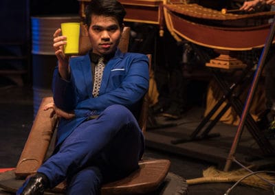 Phare Circus performance "Khmer Metal": bar patron in suit and tie sits in makeshift chair on a tire holding a drink
